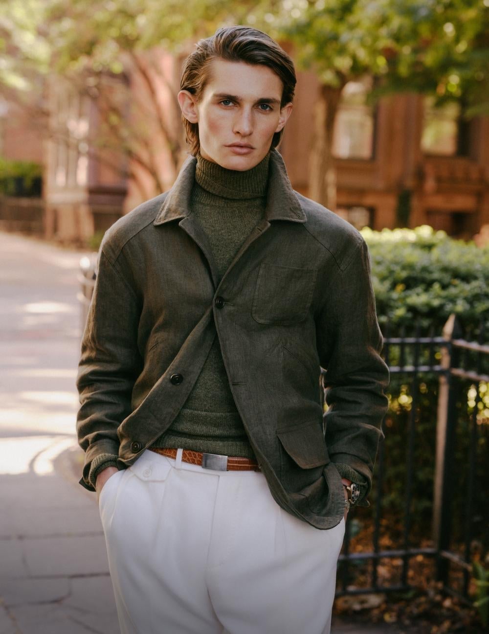 Man leaning against wall wears brown jacket and white pants.